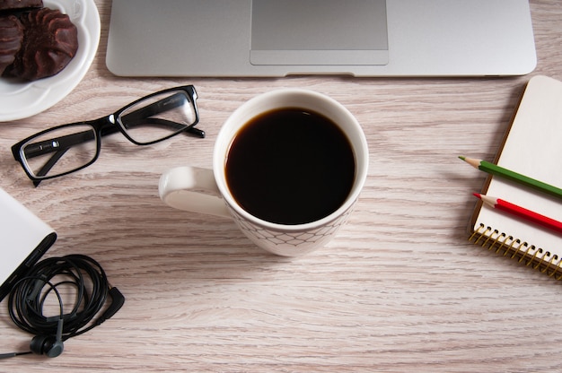 Top view of laptop and notepad with glasses and headphones on rustic wood desktop and cup of coffee.