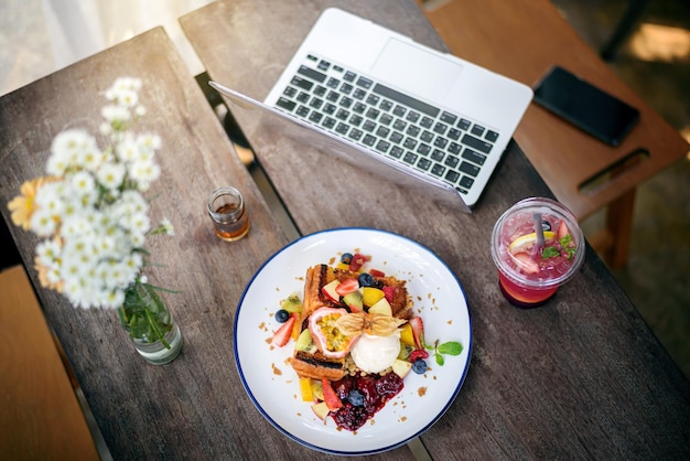 Top view of laptop and smartphone with French toast topping mixed fruit ice cream italian soda on wooden table in modern bright kitchen interior