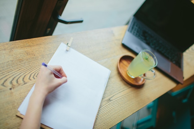 Top view of left hand of woman writing something on white paper. Girl sitting at desk in cafe. Blurred image of wooden table with laptop computer and lemon drink. Student or business woman