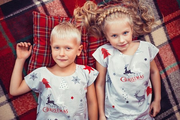 Top view of little blonde girl and boy in christmas pajamas lying on checkered blanket and pillows