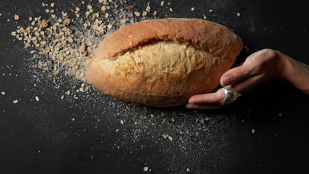 Top view of loaf of fresh bread with crumbs and flour over black background. Hot fresh bread after baking. Woman's hand touching bread. Bakery concept.