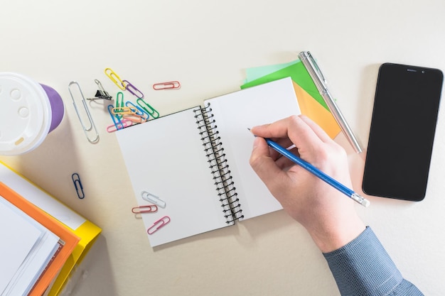 Top view of a male hand writing in a notebook on a office desk with coffee cup folders supplies office and smart phone