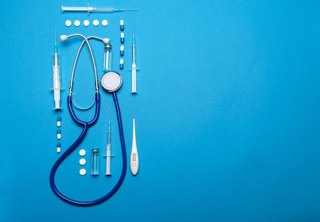 Top view of medical supplies - stethoscope, pills, syringe, thermometer and vaccine ampoule on blue background