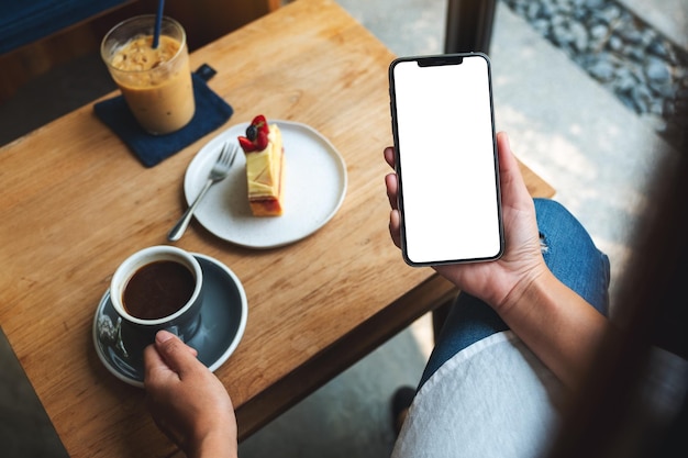 Top view mockup image of a woman holding mobile phone with blank white desktop screen while drinking coffee in cafe