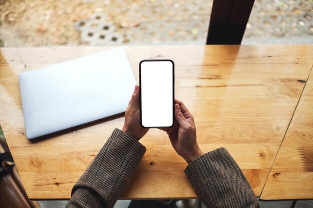 Top view mockup image of a woman holding mobile phone with blank white desktop screen with laptop on table