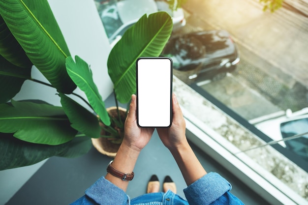 Top view mockup image of a woman standing and holding black mobile phone with blank white screen