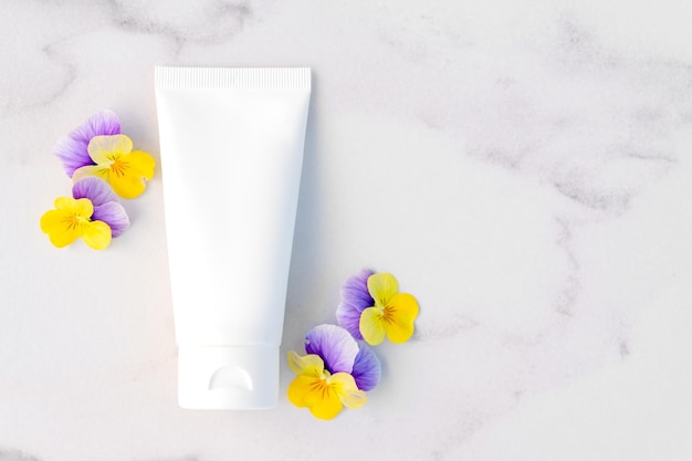 Top view of mockup of white squeeze bottle plastic tube for branding of cosmetics cream gel skin care Cosmetic bottle container on marble table with copy space and pansy flowers