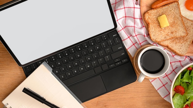 Top view of a modern workspace with digital tablet computer, notebook, coffee cup and breakfast set.