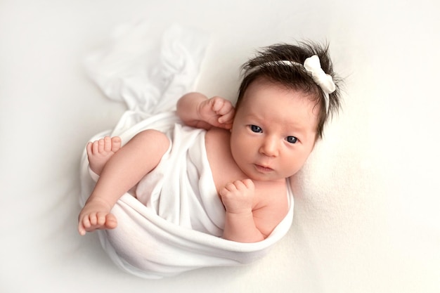 Top view of a newborn baby girl lying in a white cocoon on a white bed with a white bandage Beautiful portrait of a little girl with wide open eyes 7 days one week Photography in macro studio