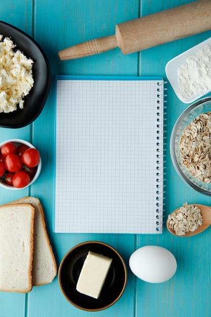 Top view of notepad with cottage cheese tomatoes butter bread slices egg flour oats and rolling pin around on blue background