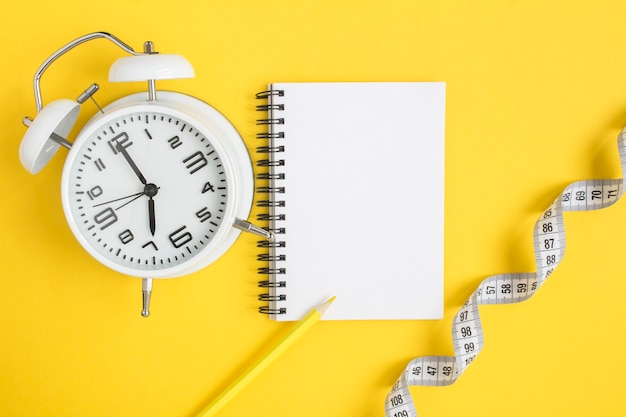 Photo top view of open notepad, white alarm clock and white centimeter on yellow, flat lay