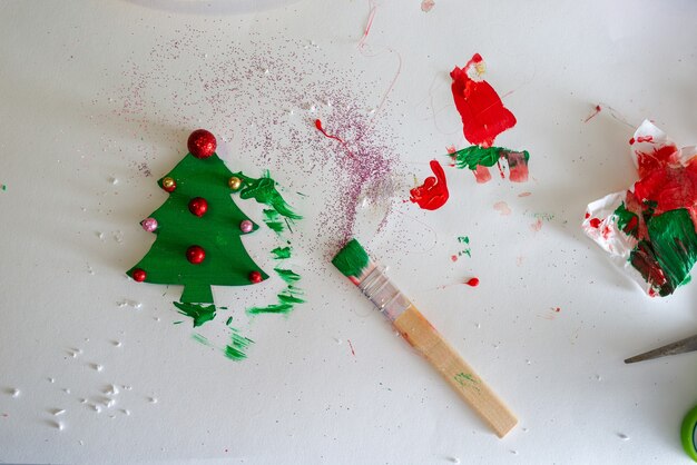 Top view of a paintbrush on a working surface next to a handmade and colored wooden christmas tree ornament.