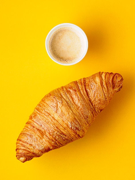 Top view on paper cup of coffee and croissant on yellow background