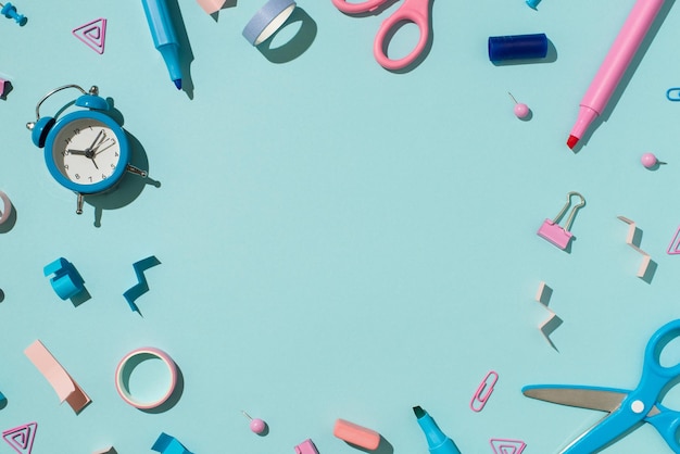 Top view photo of bicolor blue and pink school supplies felt pens binder clips pins eraser adhesive tapes scissors and alarm clock on isolated pastel blue background with empty space in the middle