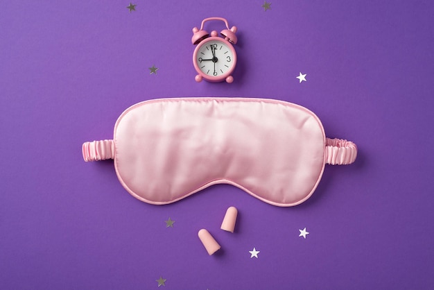 Top view photo of the big silk sleep mask and small pink clock above it with scattered confetti in shape of stars and two pink earplugs on the violet background