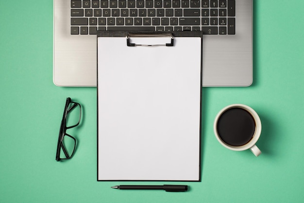 Top view photo of black clipboard folder with white paper sheet on laptop keyboard pen glasses and cup of coffee on isolated turquoise background with copyspace