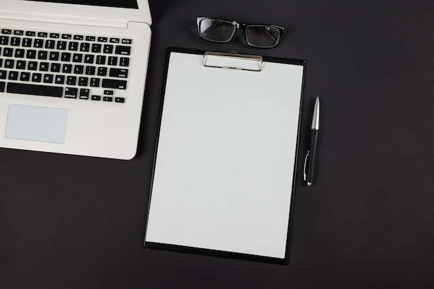 Top view photo of black clipboard folder with white paper sheet pen glasses and laptop on isolated black background with blank space