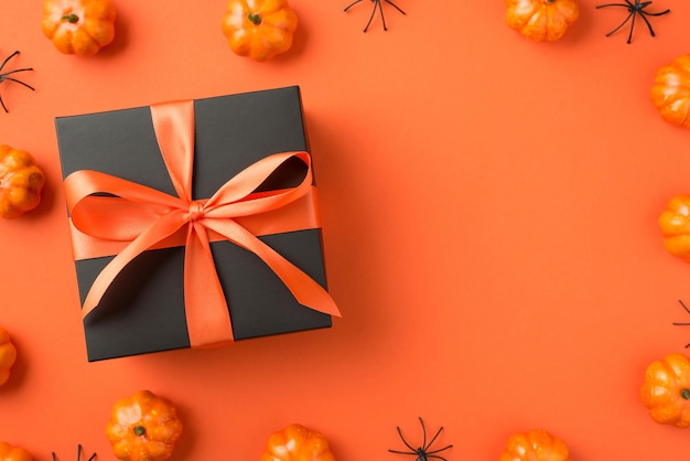 Top view photo of black giftbox with orange ribbon bow small pumpkins and spiders on isolated orange background with blank space