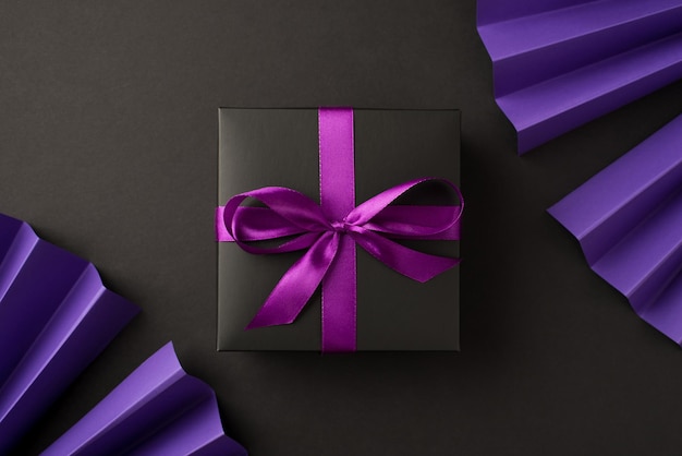 Top view photo of black giftbox with purple ribbon bow and violet paper fans on isolated black background