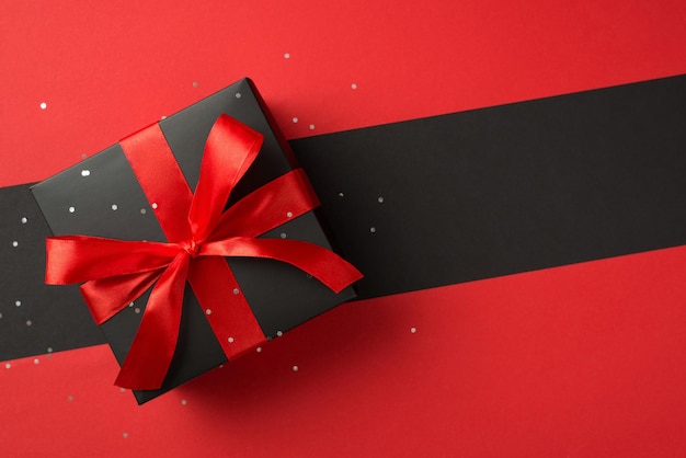 Top view photo of black giftbox with vivid red ribbon bow and confetti on isolated bicolor red and black background with copyspace