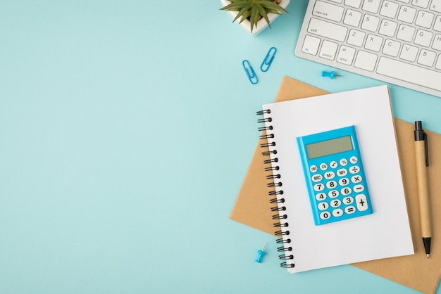 Top view photo of blue calculator on copybooks pen pins clips plant and keyboard on isolated pastel blue background with copyspace