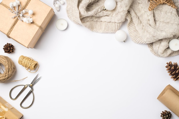 Top view photo of craft paper giftbox with snow twig sweater christmas tree balls handicraft tools scissors spools of twine candles and cones on isolated white background with copyspace