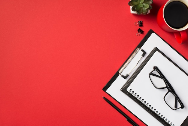 Top view photo of cup of drink flowerpot two binder clips black pen clipboard folder with paper sheet and glasses on two planners on isolated vivid red background with blank space