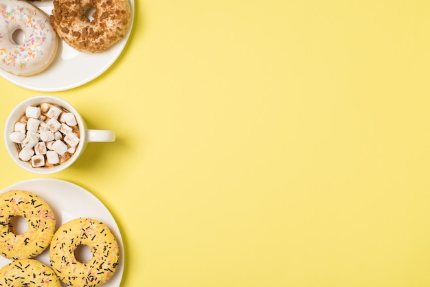 Top view photo of cup of drink with marshmallow and two plates with different color glazed donuts on isolated light yellow background with copyspace