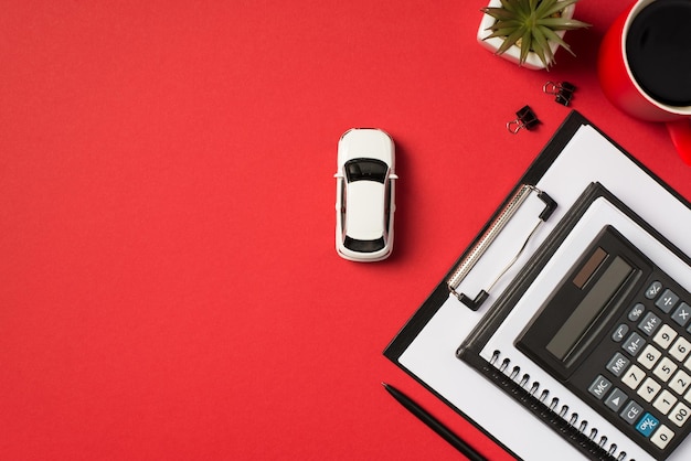 Top view photo of cup of white car model in the middle coffee flowerpot binder clips pen black clipboard folder two notepads and calculator on isolated vivid red background with empty space