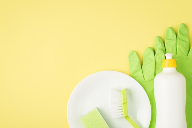 Top view photo of detergent foam bottle without label green sponge and brush on dish rubber gloves on isolated yellow background with copyspace