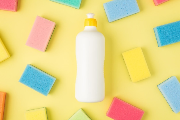 Top view photo of detergent white container without label and multicolor sponges on isolated yellow background with blank space