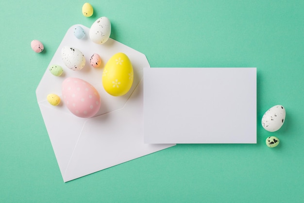 Top view photo of easter decorations white open envelope multicolored easter eggs and paper sheet on isolated turquoise background with copyspace