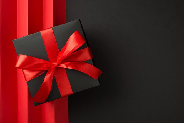 Top view photo of giftbox in black packaging with vivid red ribbon bow and red sheet with vertical folds on isolated black background with copyspace