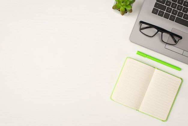 Top view photo of glasses on grey laptop plant light green open diary and pen on isolated white wooden table background with empty space