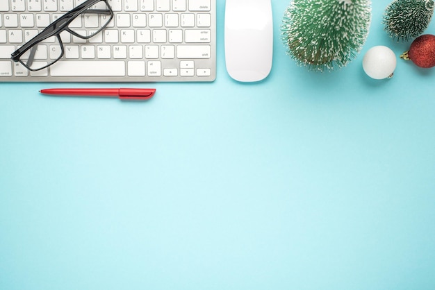 Top view photo of glasses on white keyboard computer mouse pen pine toys red and white christmas tree balls on isolated pastel blue background with copyspace