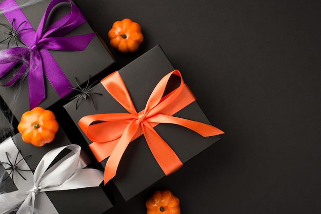 Top view photo of halloween composition three black gift boxes with violet orange and white ribbon bows small pumpkins spiders and web on isolated black background with copyspace