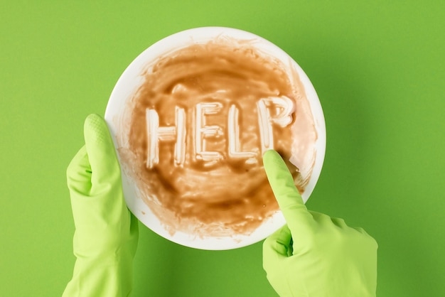 Top view photo of hands in green rubber gloves holding dirty plate and drawing word help with forefinger on isolated green background