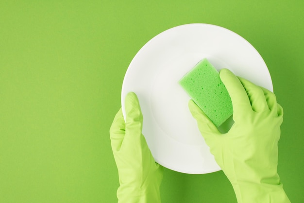 Top view photo of hands in green rubber gloves holding white plate and using green scouring pad on isolated green background with blank space