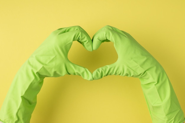 Top view photo of hands in green rubber gloves making heart with fingers on isolated yellow background with copyspace