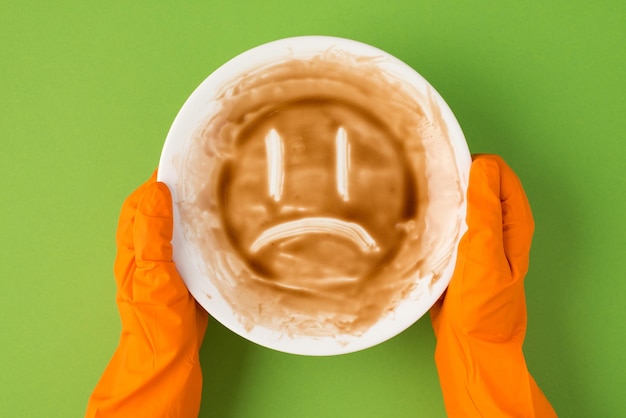 Top view photo of hands in orange rubber gloves holding dirty plate with drawn sad face on isolated green background