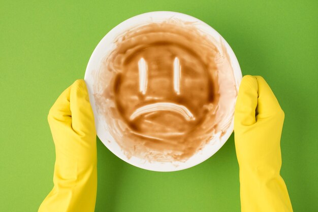 Top view photo of hands in yellow rubber gloves holding dirty dish with drawn sad face on isolated green background