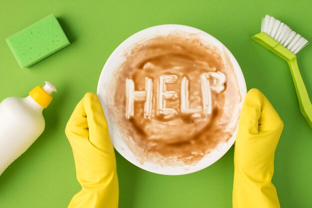 Top view photo of hands in yellow rubber gloves holding dirty dish with inscription help white detergent gel bottle green brush and scouring pad on isolated green background