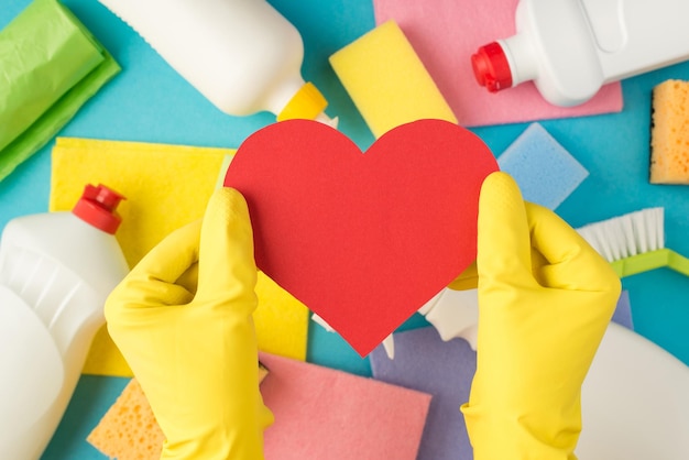 Top view photo of hands in yellow rubber gloves holding red paper heart multicolor sponges viscose rags garbage bags brush and detergent bottles on isolated pastel blue background with blank space