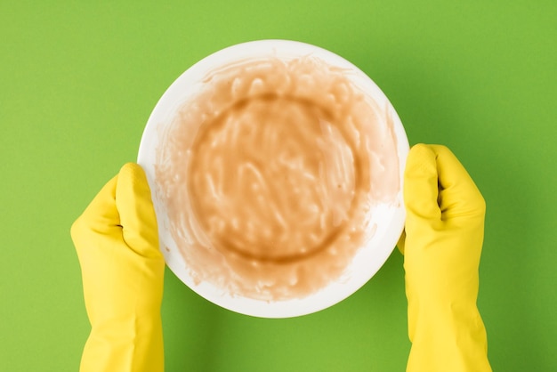 Top view photo of hands in yellow rubber gloves holding unclean plate on isolated green background