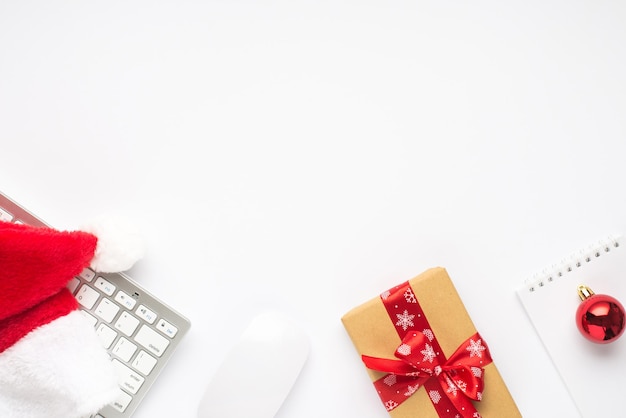 Top view photo of keyboard computer mouse santa claus hat red christmas tree ball notepad and gift box with red ribbon bow on isolated white background with copyspace