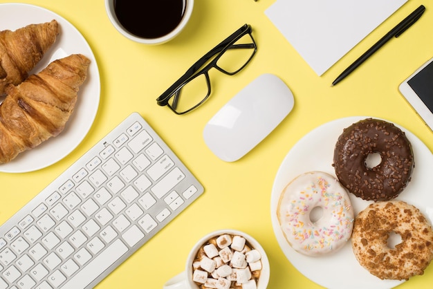 Top view photo of keyboard mouse glasses pen notebook two cups of drink with marshmallow and coffee two plates with croissants and donuts and smartphone on isolated pastel yellow background