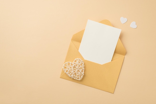 Top view photo of open pastel yellow envelope with paper sheet white hearts and rattan heart on isolated beige background with blank space