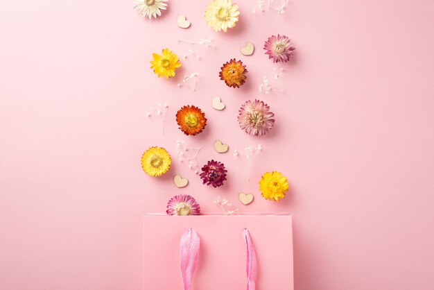 Top view photo of the paper shopping pink bag many colorful flowers with branches of gypsophila and confetti in shape of hearts scattered above bag on the pastel pink isolated empty background