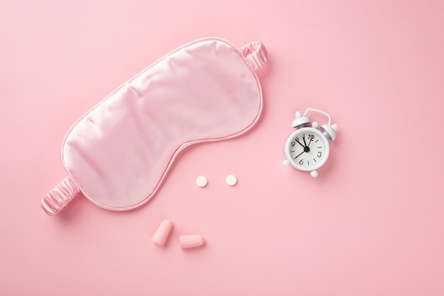 Top view photo of pink silk sleeping mask small white alarm clock pills and earplugs on isolated pastel pink background with copyspace