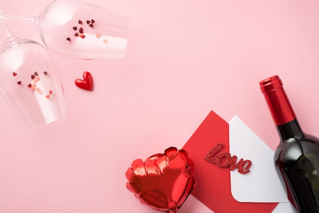 Top view photo of st valentine's day decor two wineglasses with confetti heart shaped balloon envelope red card inscription love and wine bottle on isolated pastel pink background with blank space